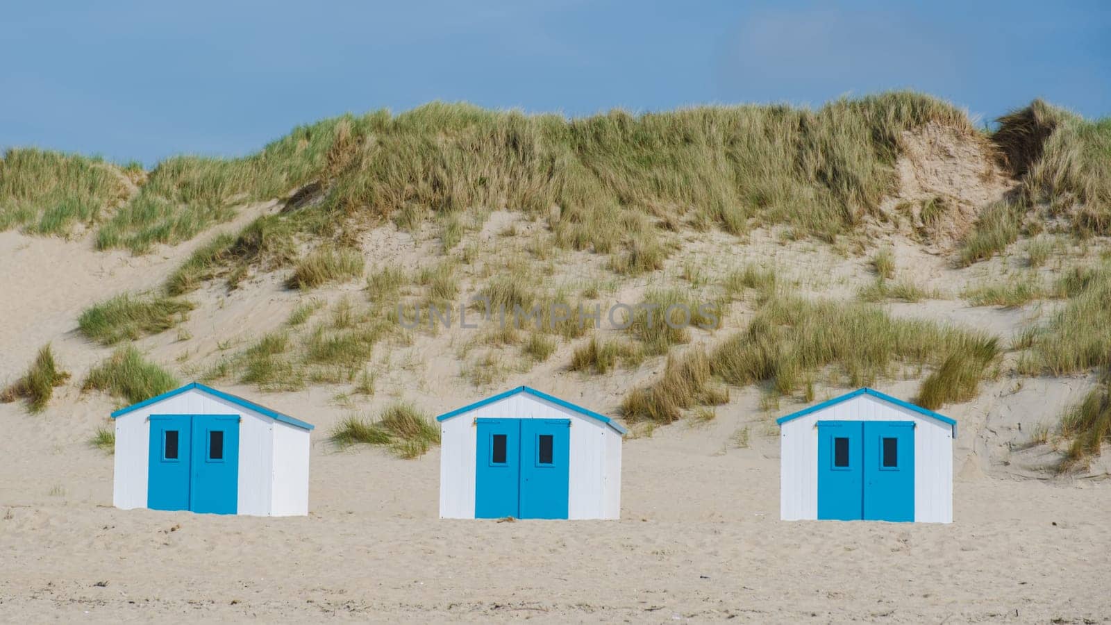 Three colorful beach huts stand in a row on the sandy shores of Texel, Netherlands, under a clear blue sky.