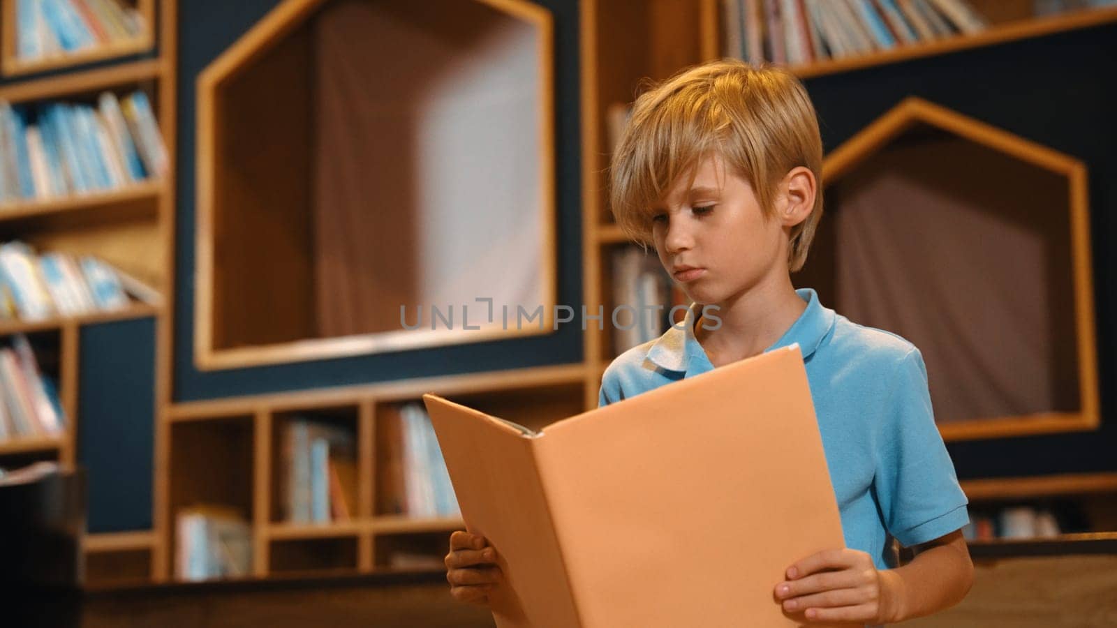 Attractive caucasian clever boy reading a book at library with stack of books placed. Smart child focus on studying, learning from novel or textbook while turning a page. Self-education. Erudition.