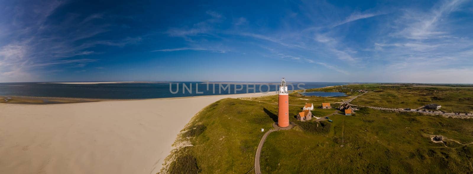 A unique perspective drone view at the lighthouse standing tall on a sandy beach, against a backdrop of the ocean waves crashing. The iconic red lighthouse of Texel Netherlands