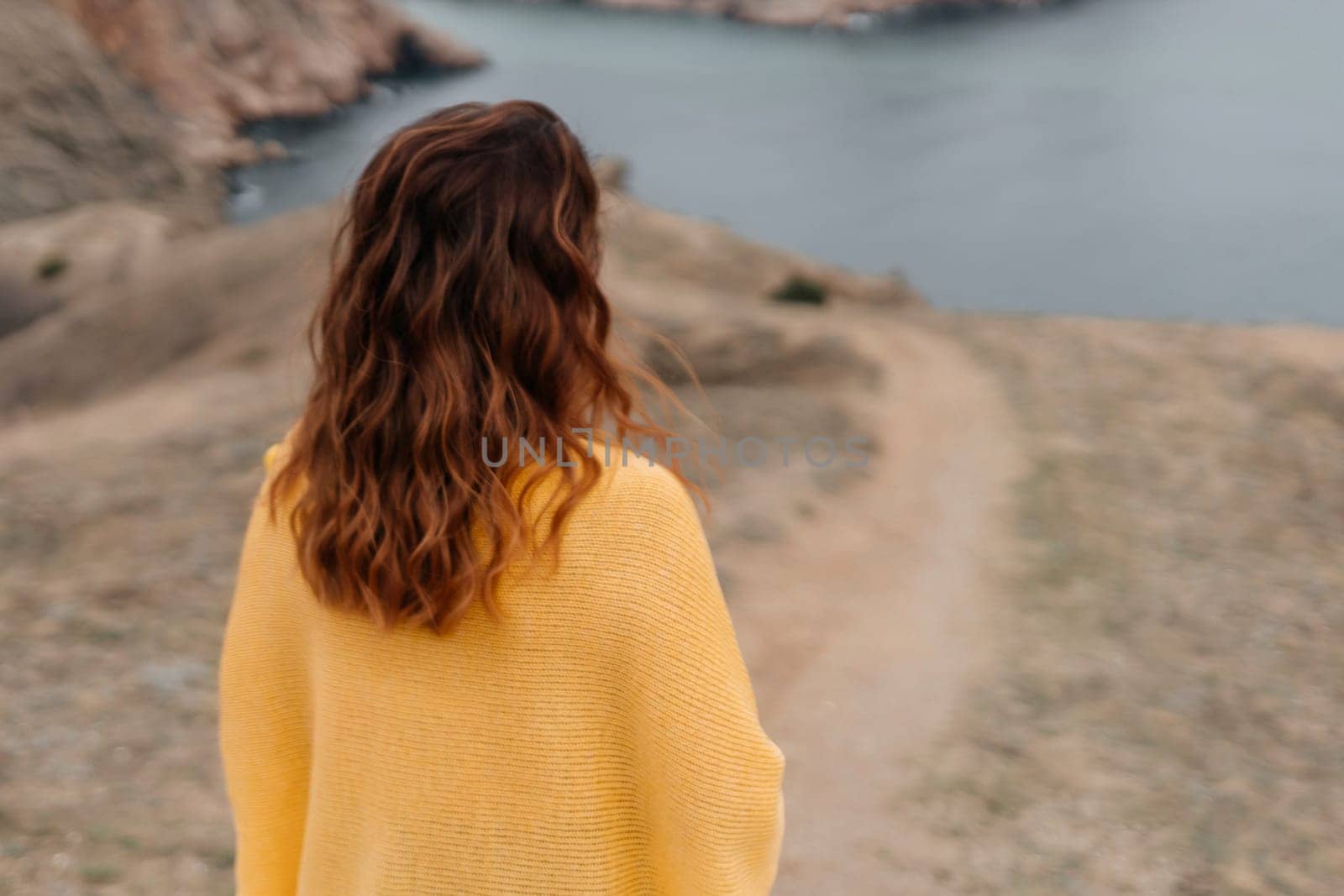 Rear view of a woman with long hair against a background of mountains and sea. Holding a bouquet of yellow tulips in her hands, wearing a yellow sweater.