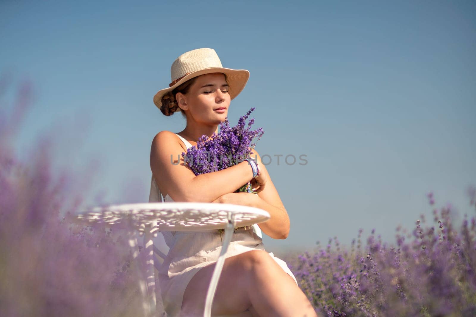 A woman is sitting in a field of lavender flowers and wearing a straw hat. She is smiling and holding a bouquet of flowers. Scene is peaceful and serene, as the woman is surrounded by the beauty of nature.