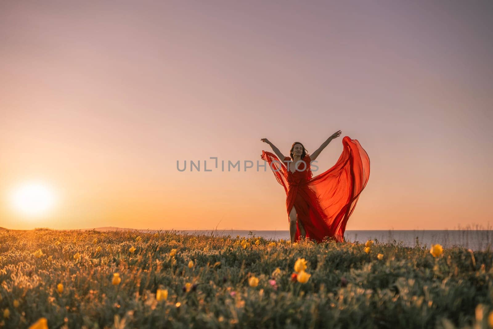 woman red dress is standing on a grassy hill overlooking the ocean. The sky is a beautiful mix of orange and pink hues, creating a serene and romantic atmosphere