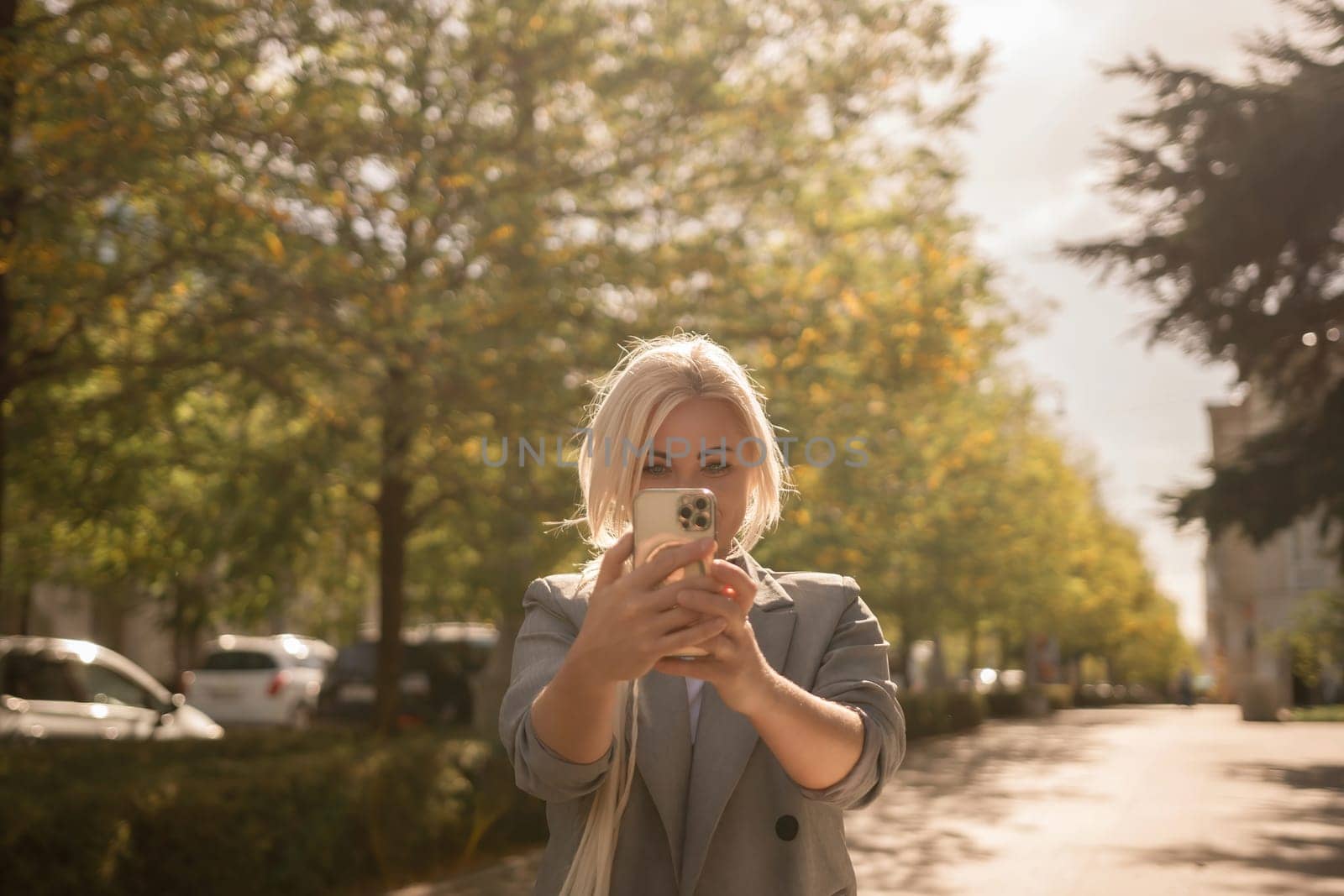 A woman is taking a picture of herself with her cell phone. She is wearing a gray jacket and scarf. The scene is set in a city with trees and cars in the background