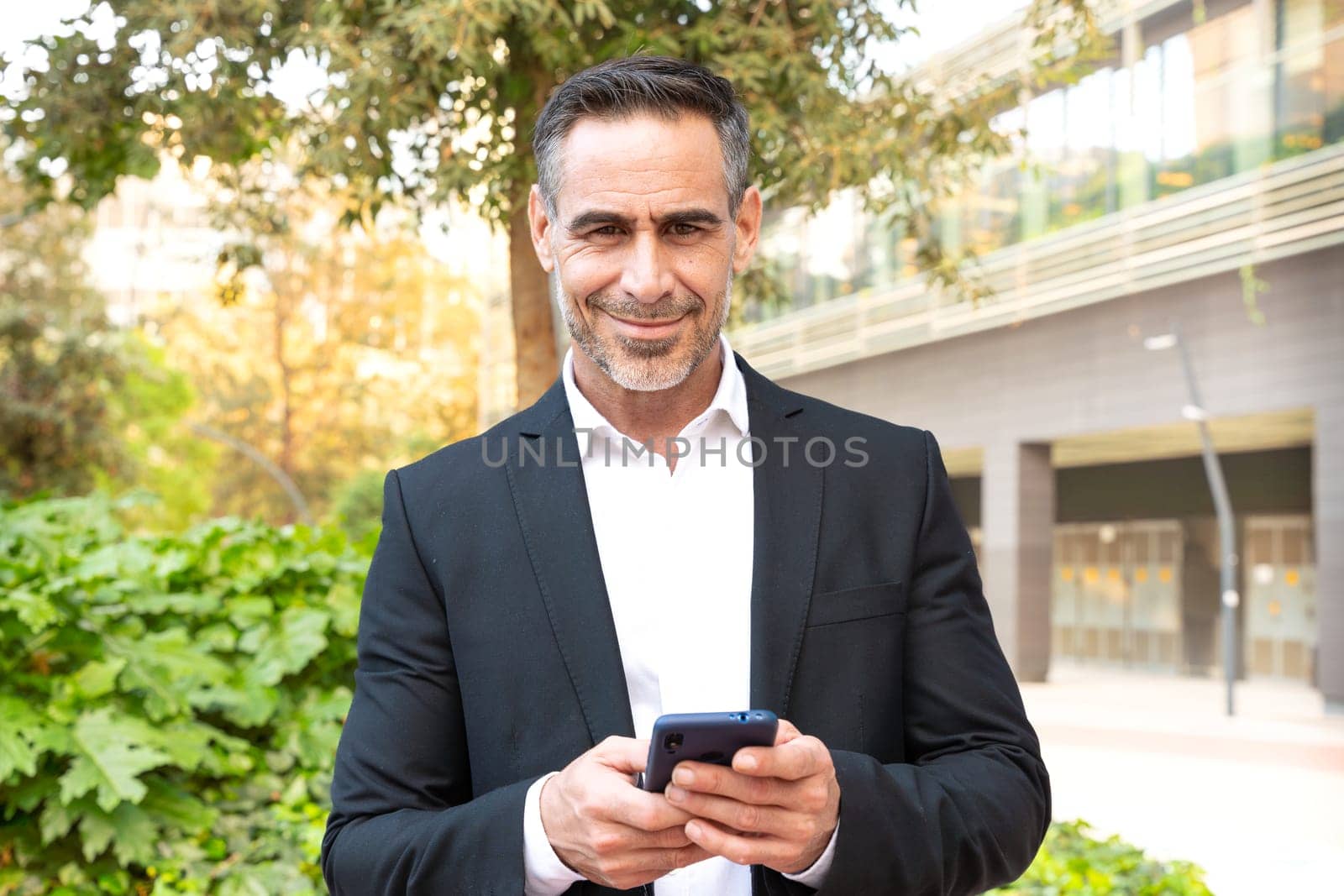 Satisfied with the results, the businessman walks down the street outside the office building, a mature boss holds a phone and coffee in his hands, writes messages and reads news online, using an app.