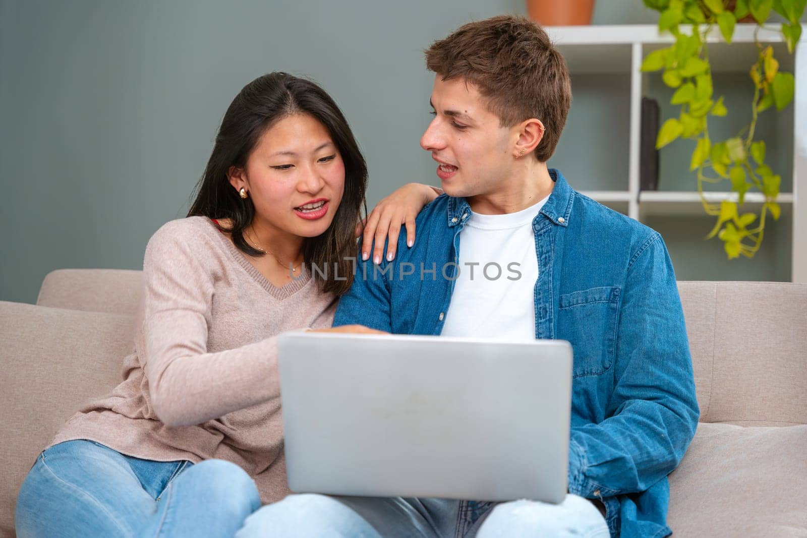 A smiling young multiracial couple sits on the couch at home using a laptop surfing the wireless internet on a gadget