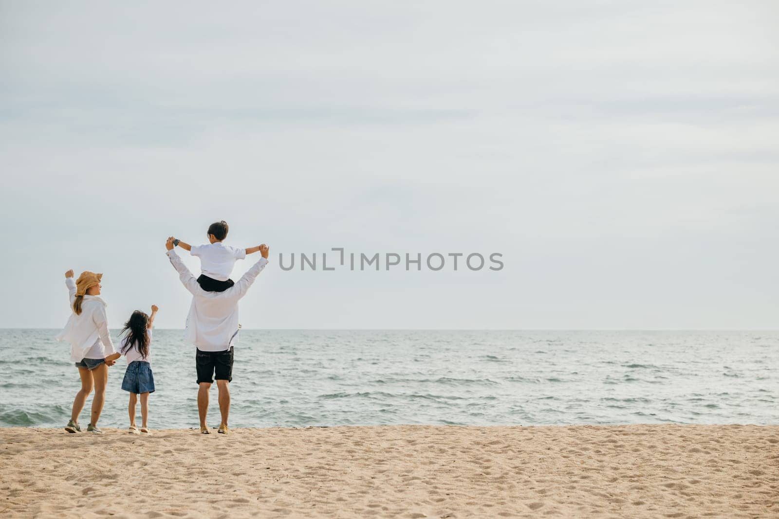 A happy family with raised arms stands on the sea beach at sunset. The father carrying his son on shoulders signifies the carefree joy and togetherness of a perfect beach vacation.