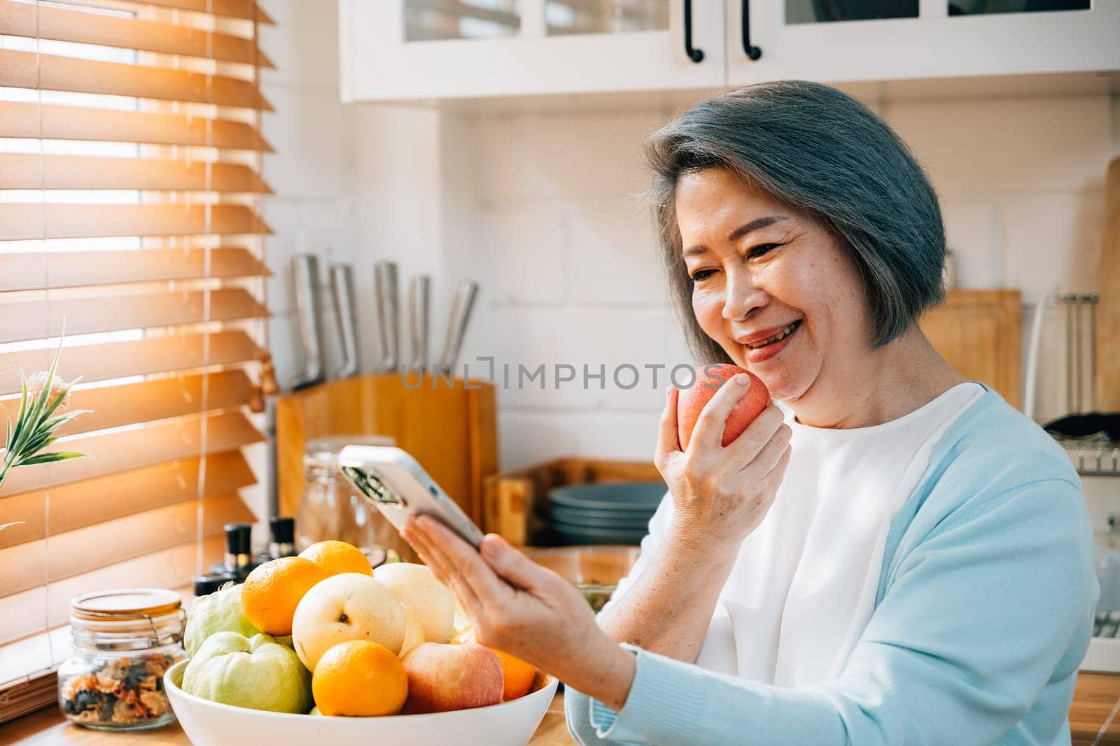 An old Asian woman, a grandmother, enjoys breakfast in the kitchen. She smiles while using her smartphone at home, eating a red apple. A portrait of modern technology and happiness.