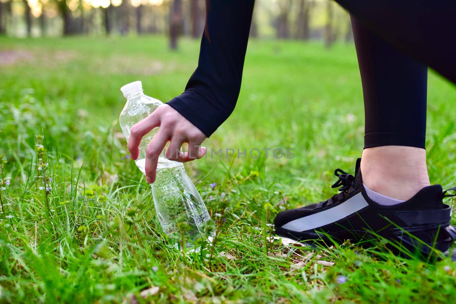 A woman's hand picks up a bottle of water that stands on the grass in the woods. healthy lifestyle