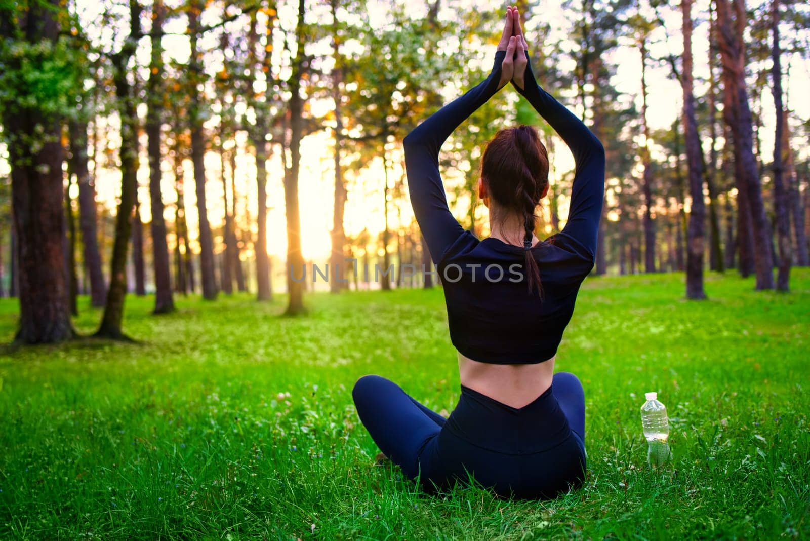 Slim beautiful woman in black tight fitting tracksuit sitting on grass in forest in yoga pose.