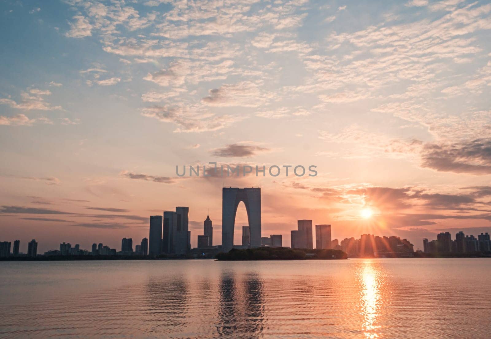 The Gate of the Orient, a distinctive skyscraper in Suzhou, China, is illuminated against the evening sky as it overlooks a calm lake. Surrounding buildings and city lights reflect on the water, creating a picturesque urban scene.