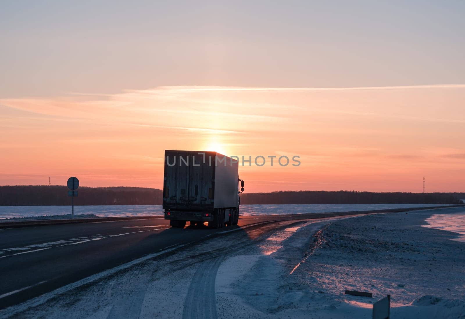 A semi truck cruises down a wintry highway as the sun sets on the horizon, casting a warm glow over the icy road and surrounding landscape.