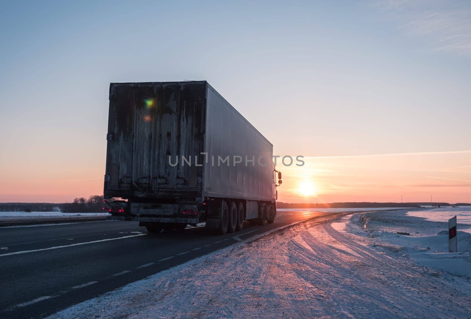 Semi truck driving along a snowy highway at sunset in winter by Busker