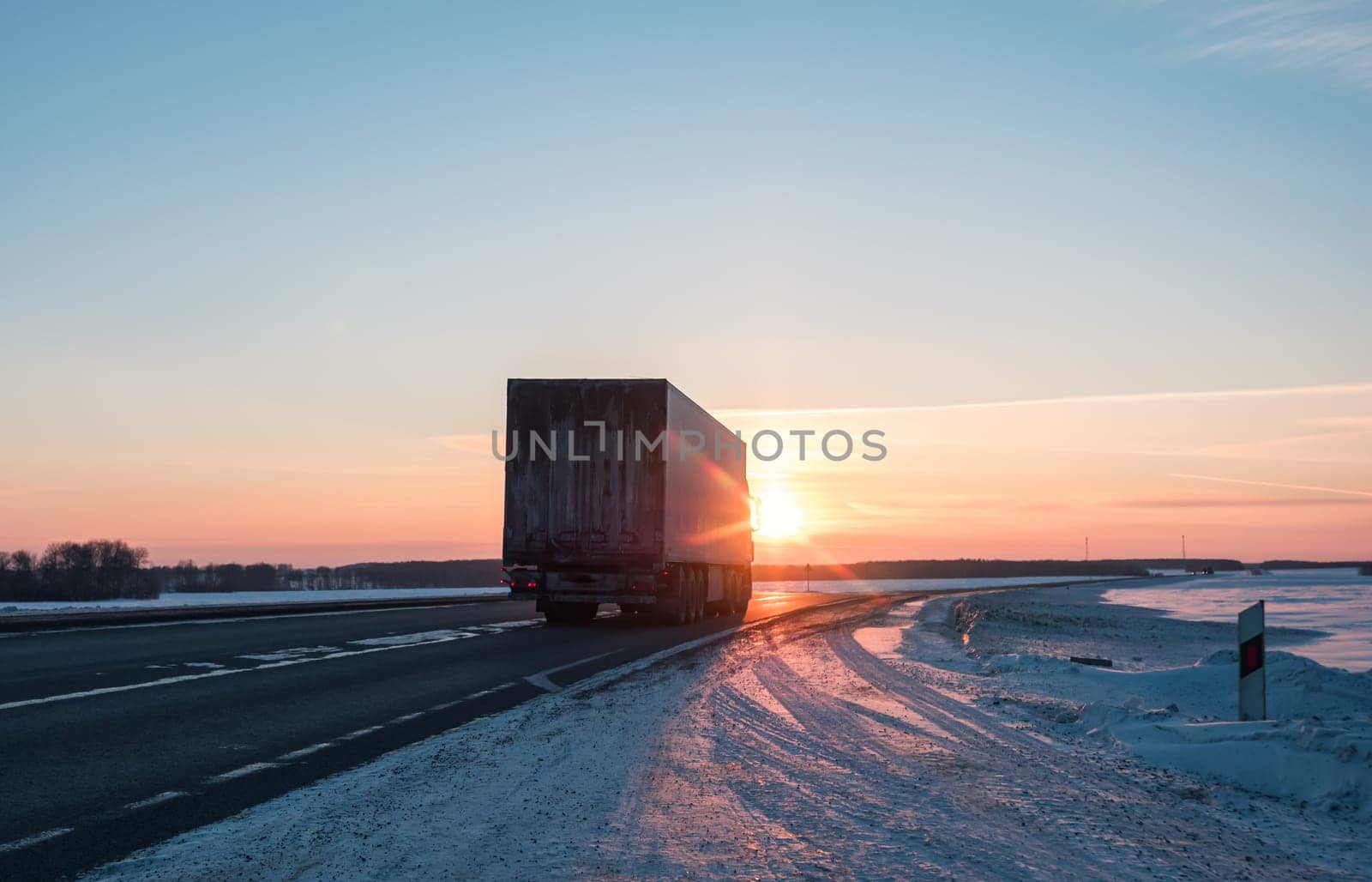 A semi truck cruises down a wintry highway as the sun sets on the horizon, casting a warm glow over the icy road and surrounding landscape.