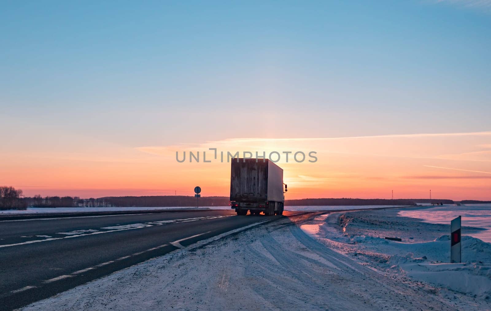 Semi truck driving along a snowy highway at sunset in winter by Busker