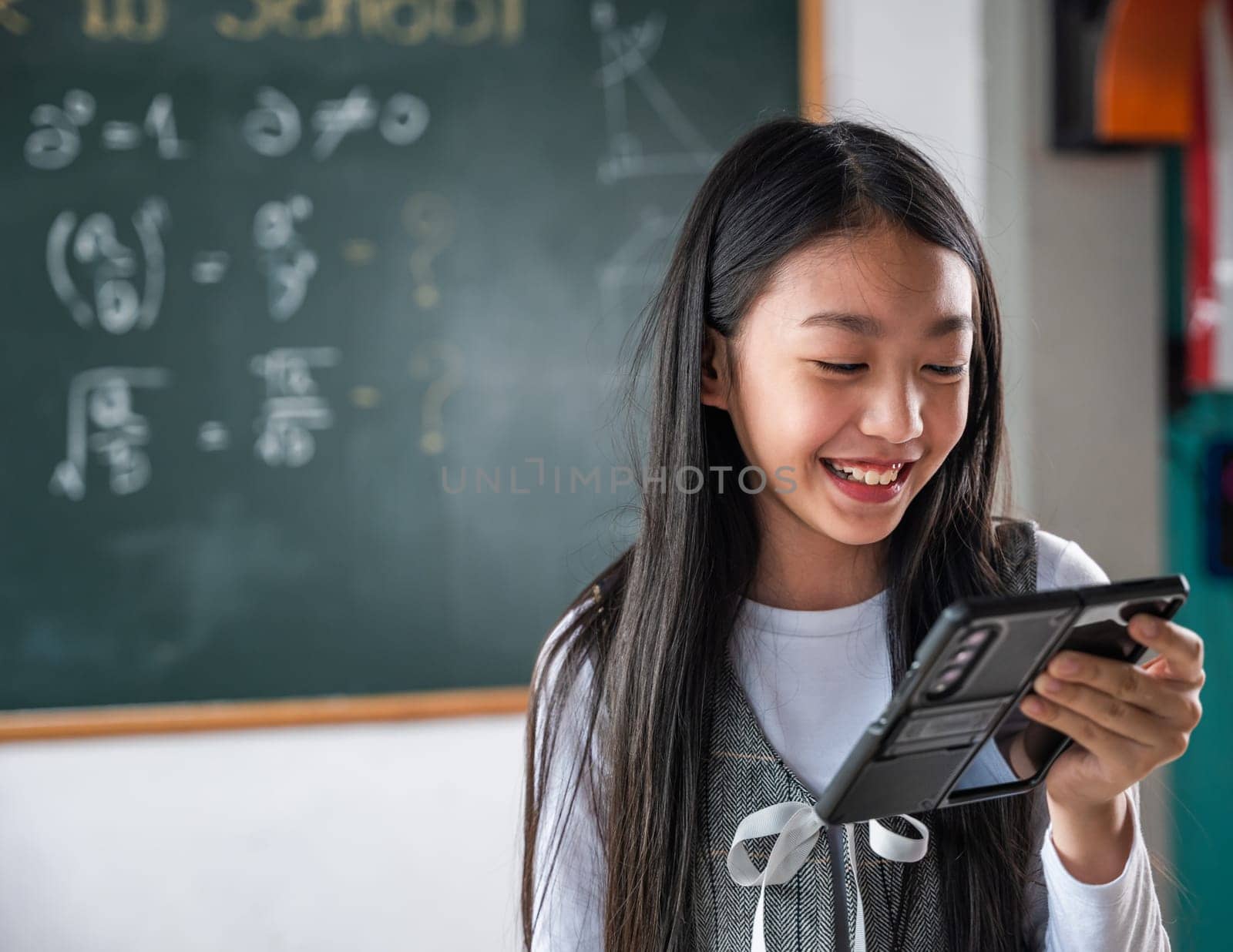 A girl is smiling while holding a cell phone in front of a blackboard with math by Sorapop