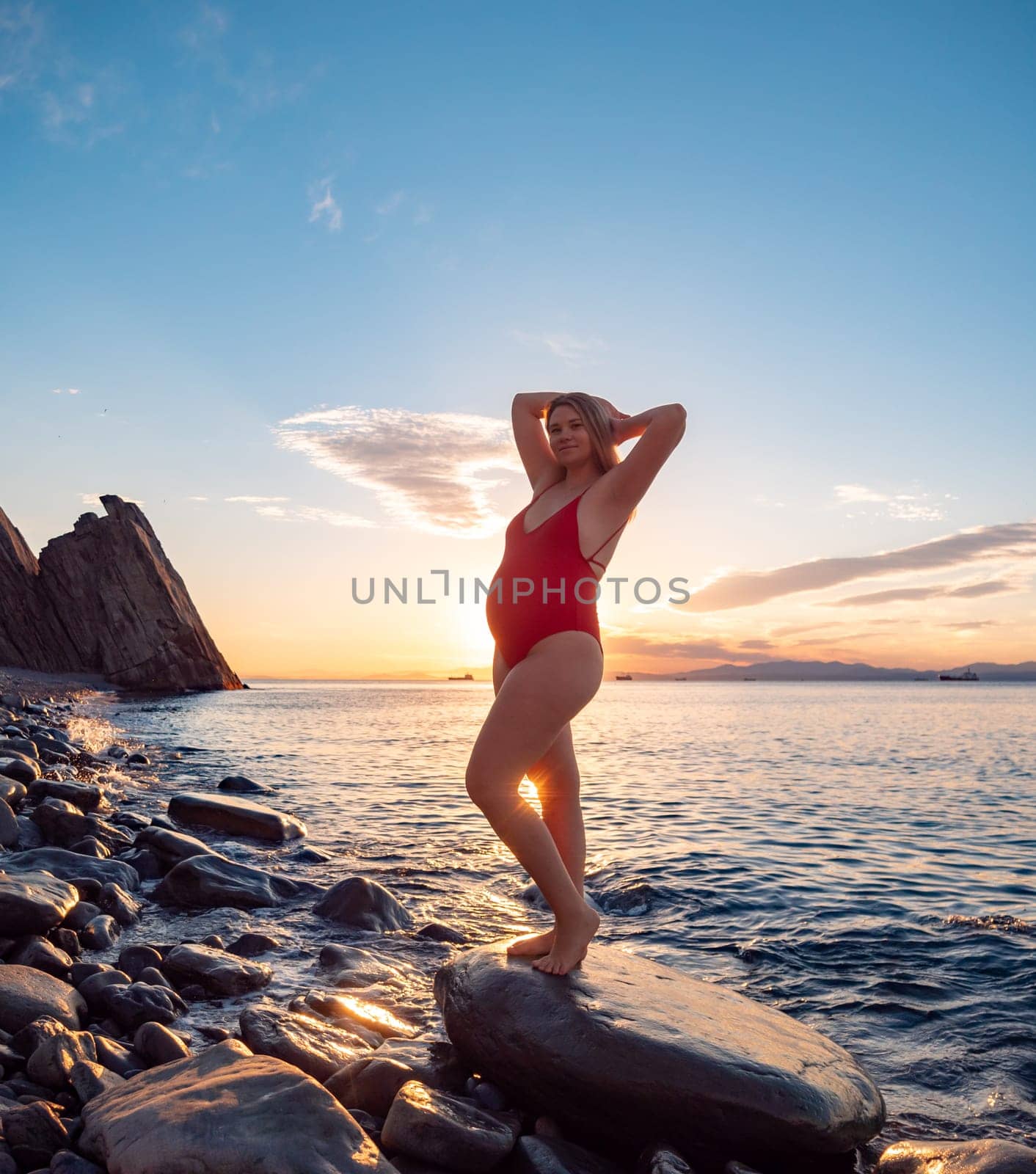 A pregnant woman stands on a rock by the seaside during sunrise.