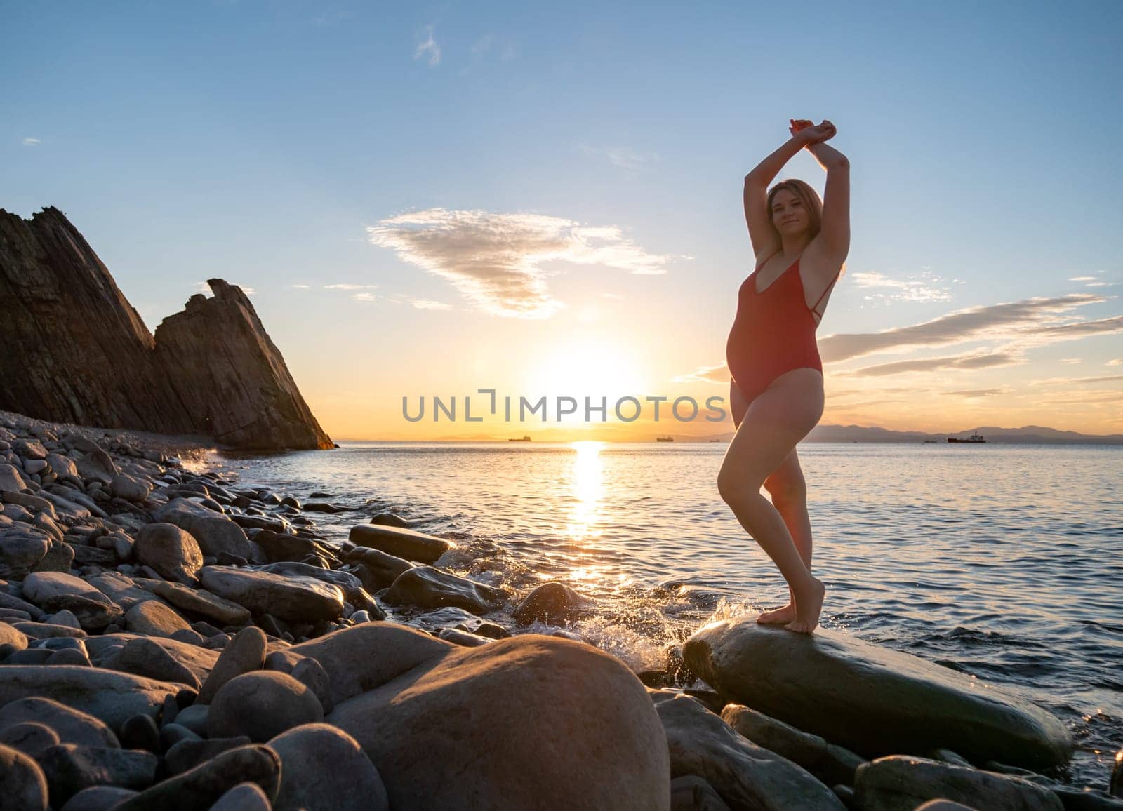 A pregnant woman stands on a rock by the seaside during sunrise.