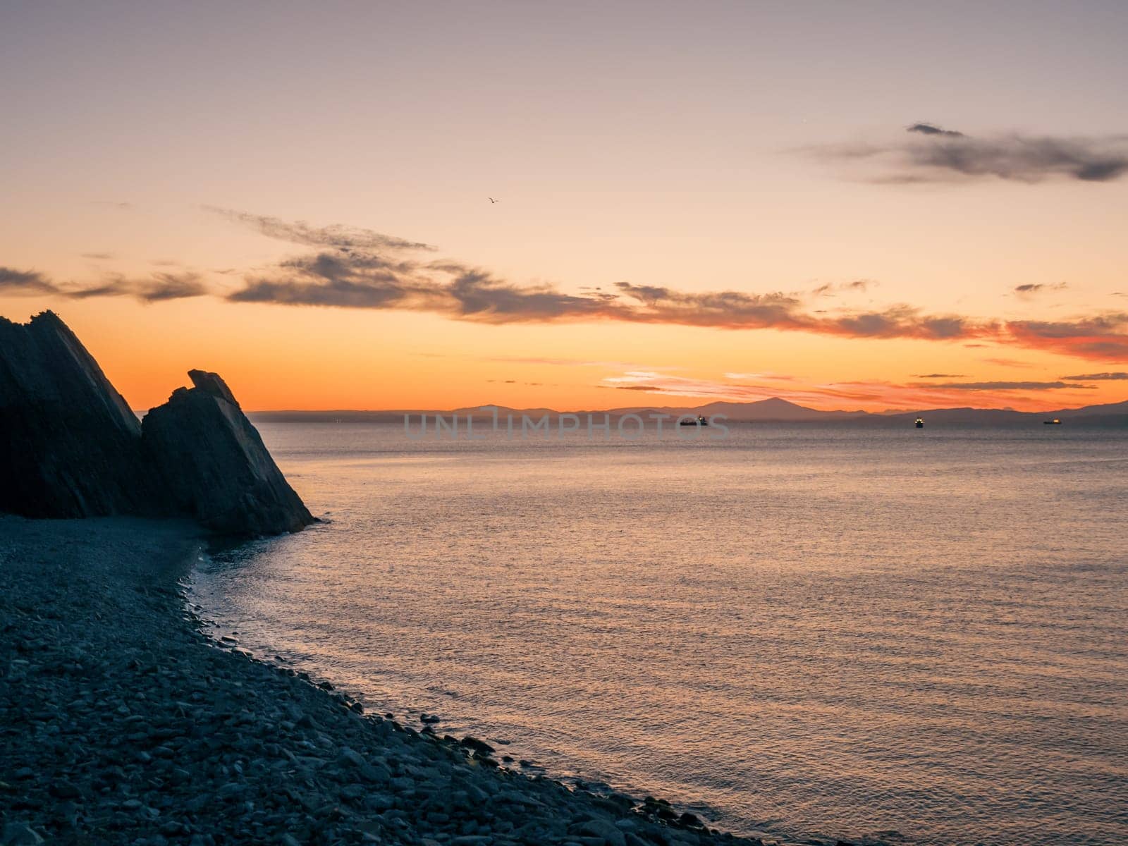 A tranquil sunset casts warm hues over a rocky beach and silhouetted cliffs. The calm ocean waters reflect the changing colors of the sky, creating a serene and picturesque coastal scene.