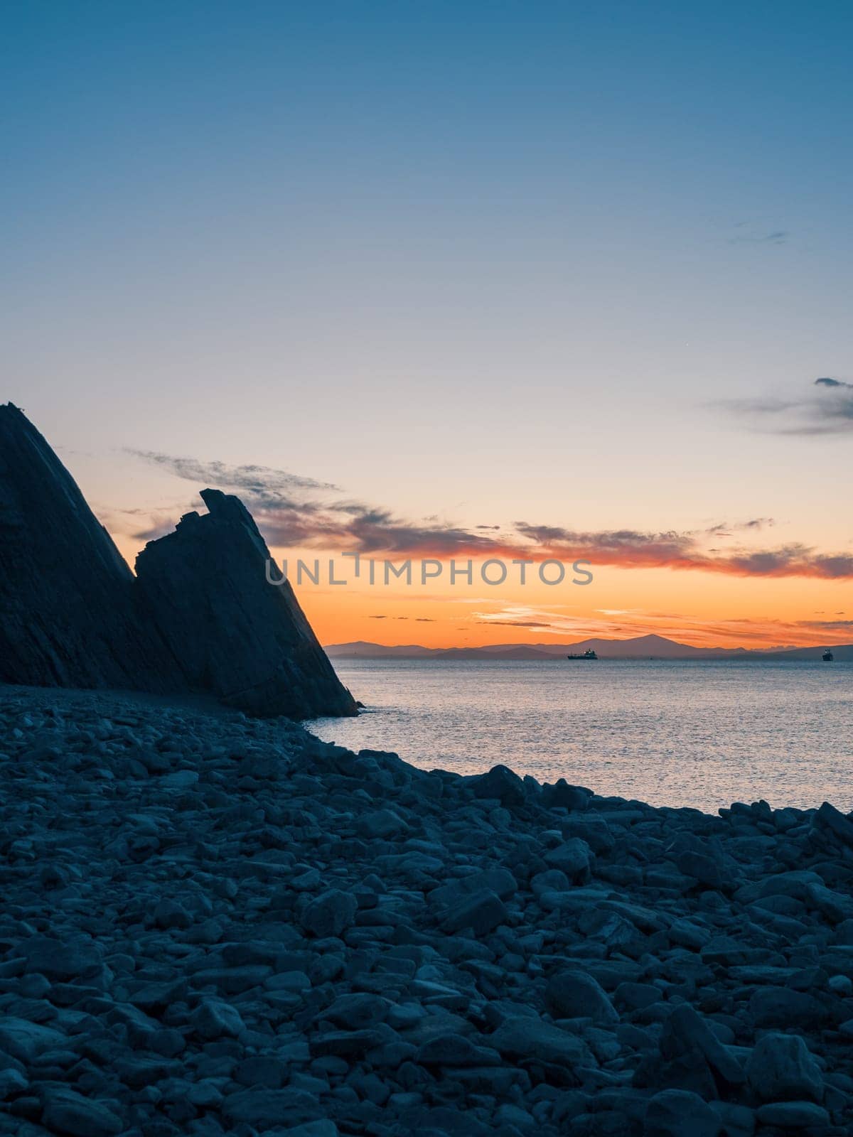 A tranquil sunset casts warm hues over a rocky beach and silhouetted cliffs. The calm ocean waters reflect the changing colors of the sky, creating a serene and picturesque coastal scene.