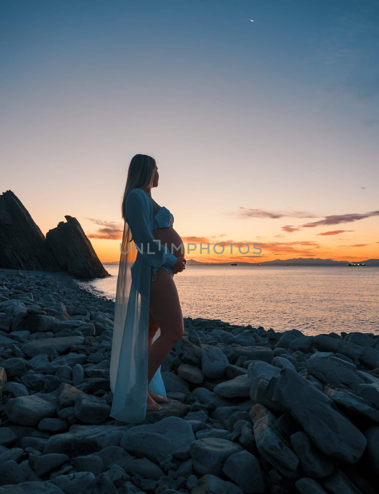 Pregnant woman in bikini posing on rocky beach at sunrise with mountain view by Busker