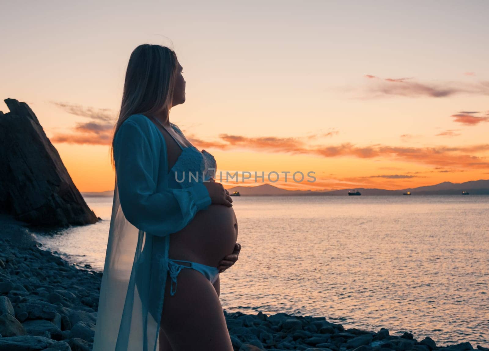 A pregnant woman in a white bikini stands on a rocky beach at sunrise, gently holding her belly. The background features calm water and a distant mountain range.