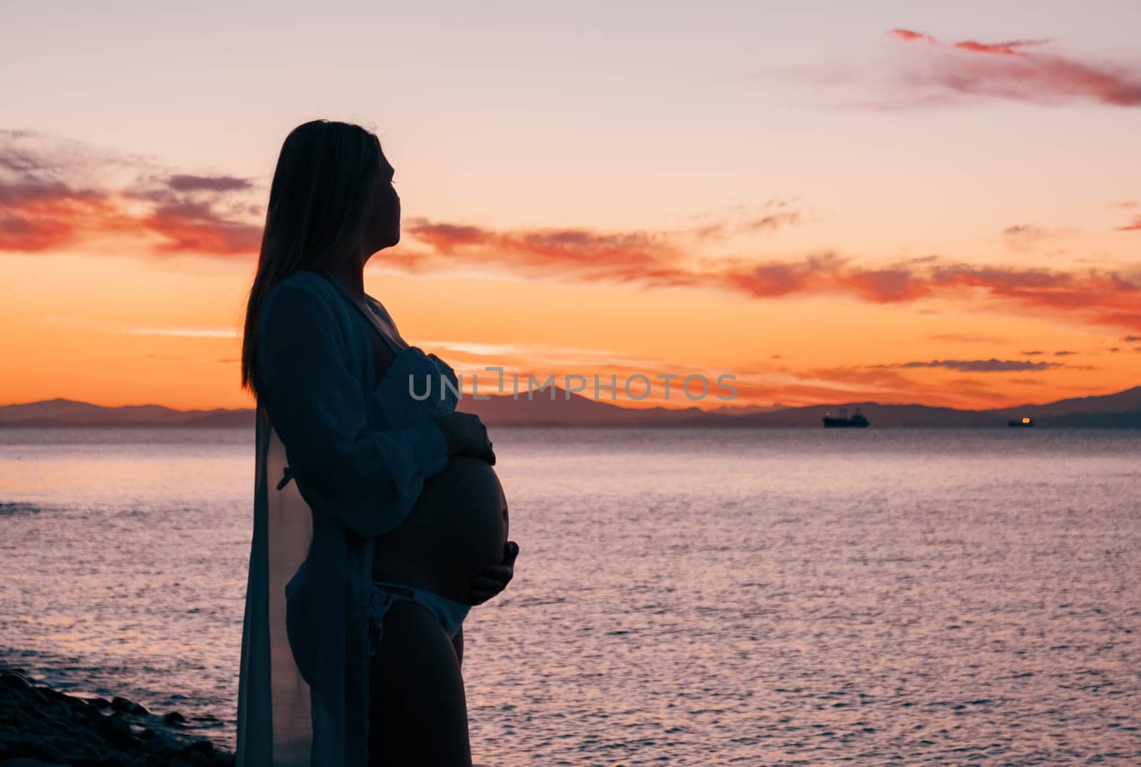 A pregnant woman in a white bikini stands on a rocky beach at sunrise, gently holding her belly. The background features calm water and a distant mountain range.