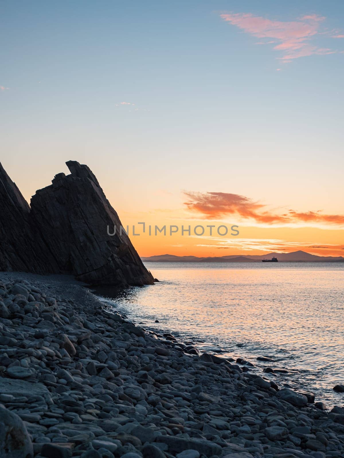 A tranquil sunset casts warm hues over a rocky beach and silhouetted cliffs. The calm ocean waters reflect the changing colors of the sky, creating a serene and picturesque coastal scene.