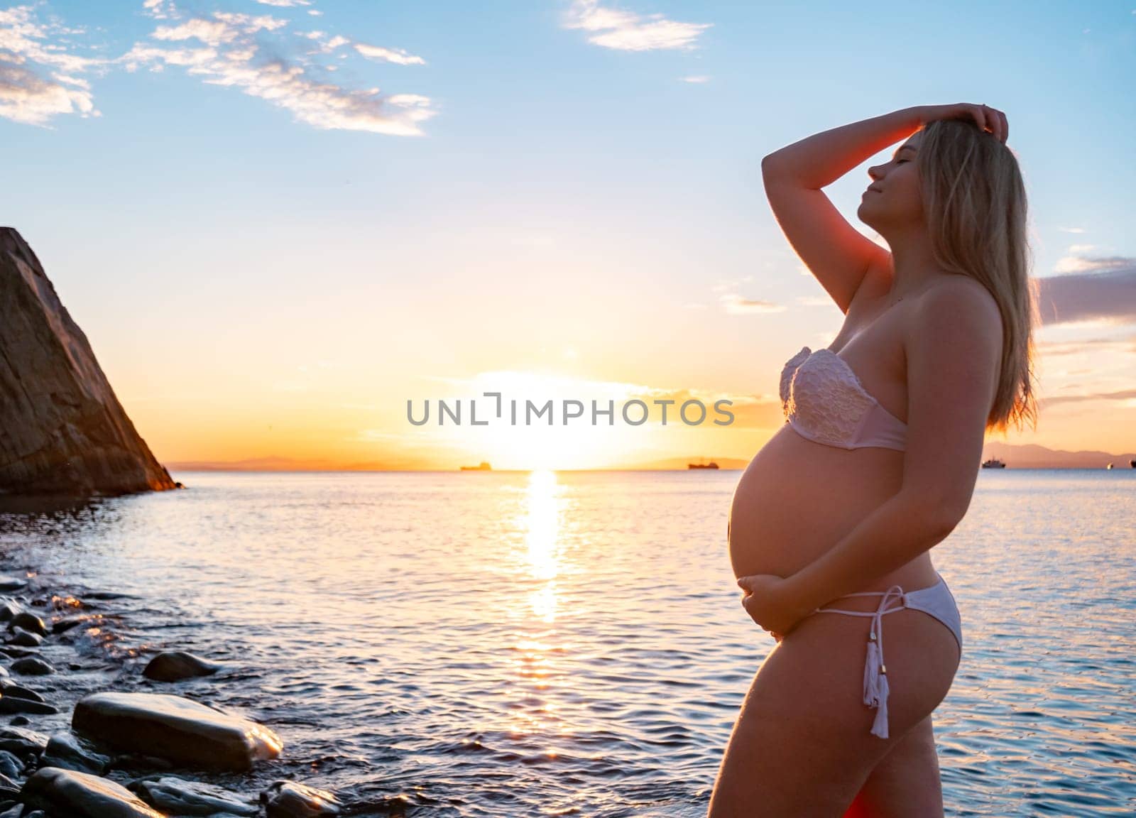 Pregnant woman in bikini posing on rocky beach at sunrise with mountain view by Busker