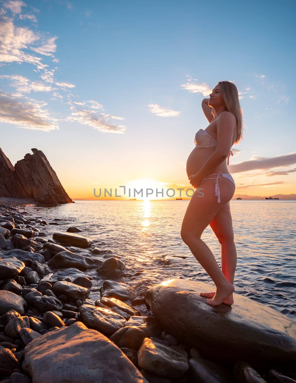 Pregnant woman in bikini posing on rocky beach at sunrise with mountain view by Busker