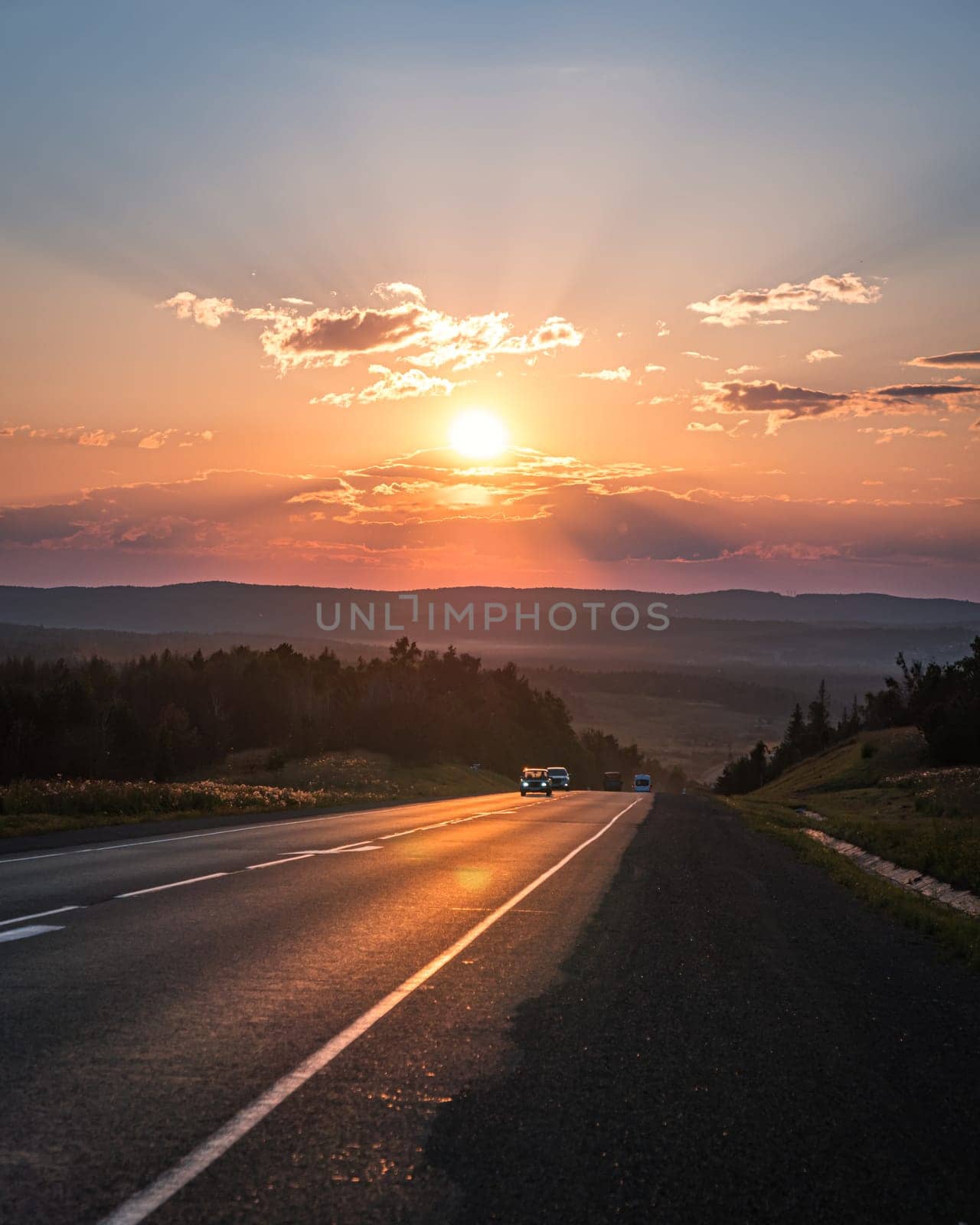 The sun sets in a fiery orange glow over a peaceful road cutting through the countryside.