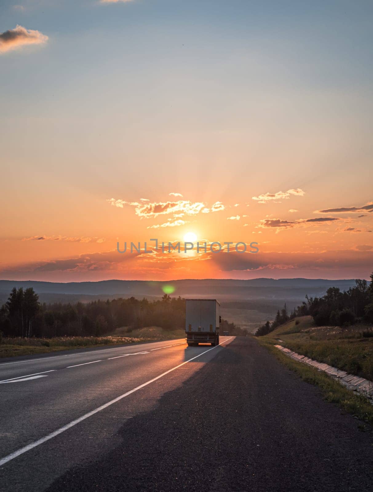 The sun sets in a fiery orange glow over a peaceful road cutting through the countryside.