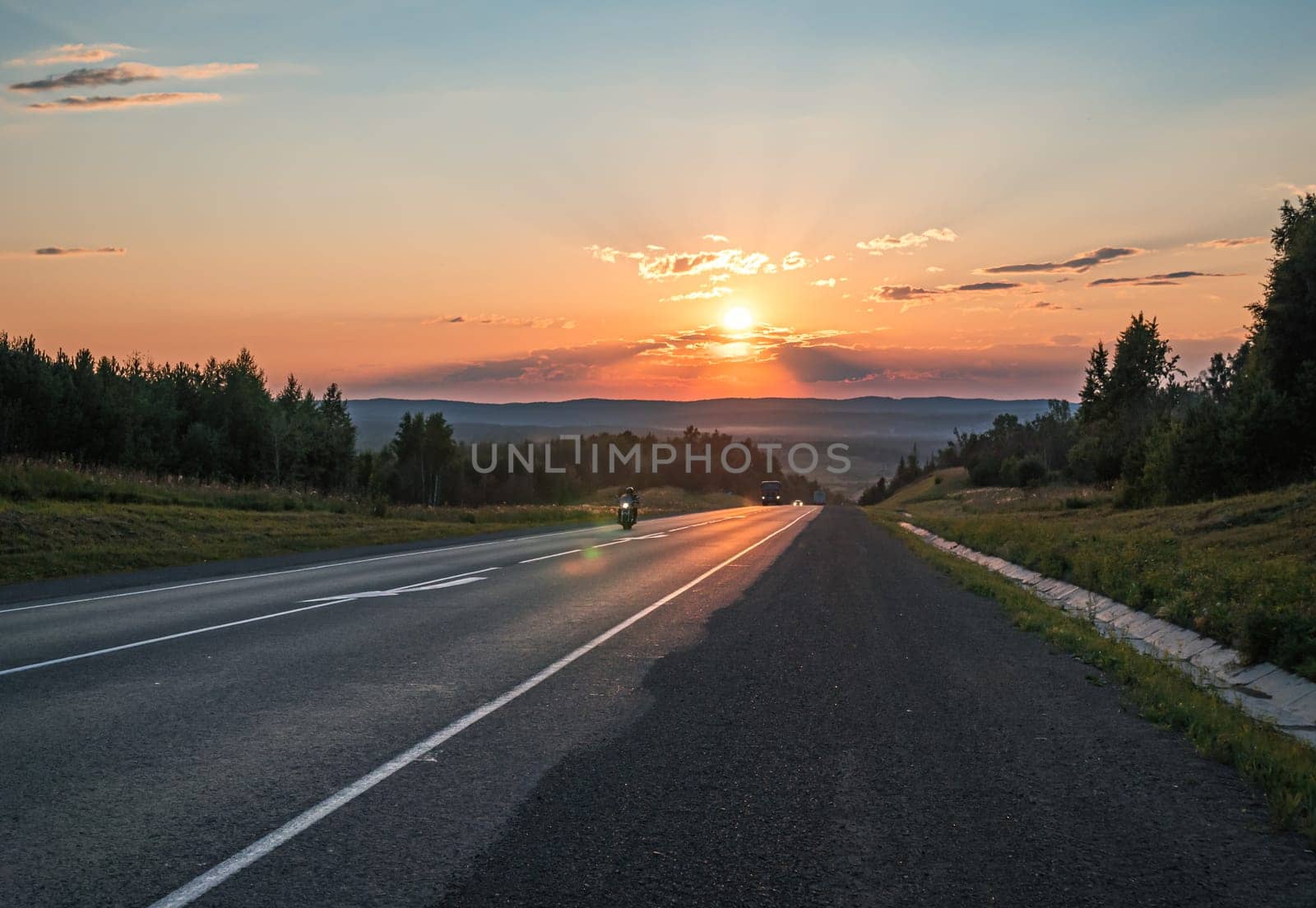 The sun sets in a fiery orange glow over a peaceful road cutting through the countryside.