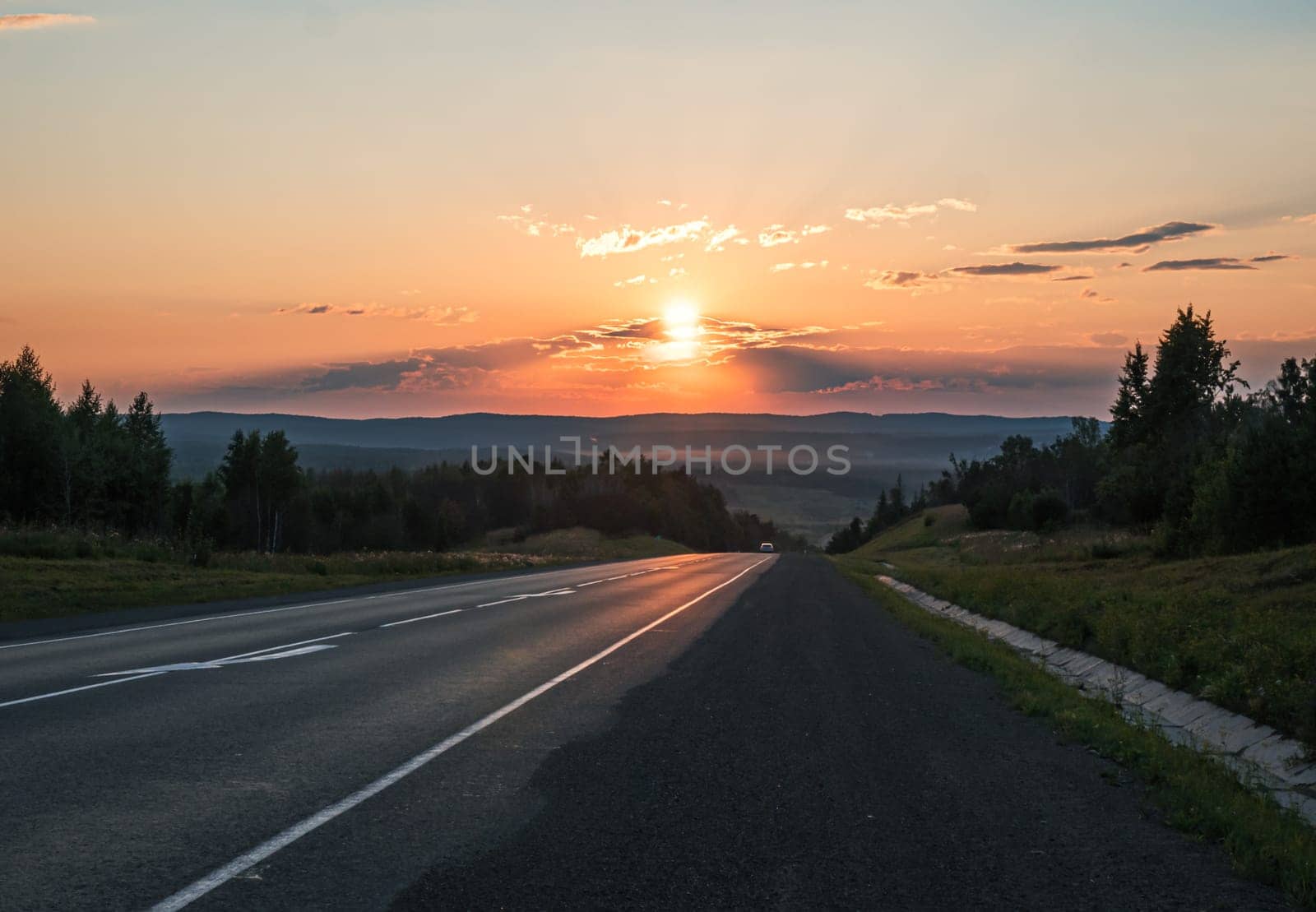 The sun sets in a fiery orange glow over a peaceful road cutting through the countryside.