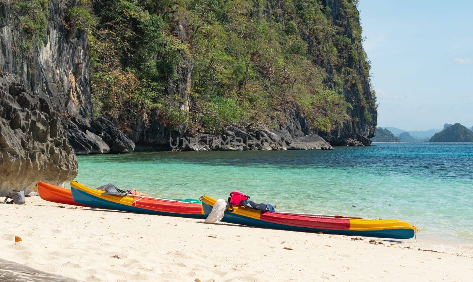 Nestled between towering rocky cliffs and turquoise waters, vibrant kayaks rest on a pristine, sandy beach under a clear, sunny sky.