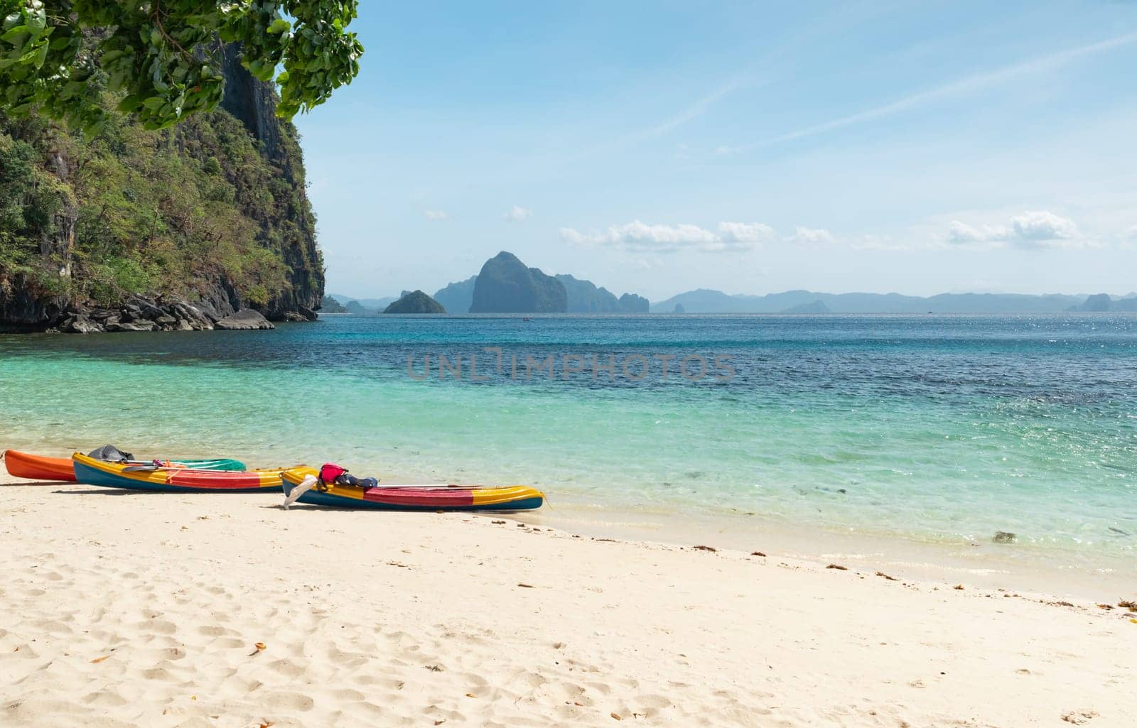 Nestled between towering rocky cliffs and turquoise waters, vibrant kayaks rest on a pristine, sandy beach under a clear, sunny sky.