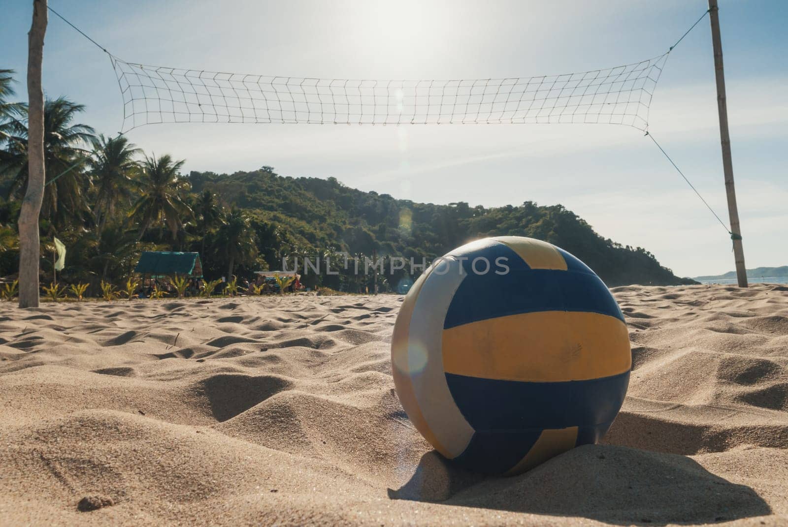 A volleyball rests on the sandy beach beneath a clear, sunny sky. A net is set up, ready for a game. Palm trees and lush green hills are visible in the background.