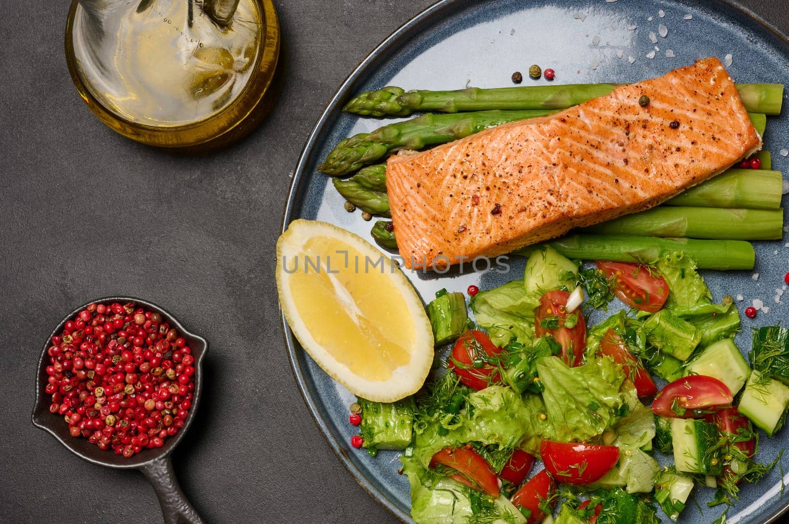 Healthy lunch with grilled salmon with asparagus and fresh tomato and cucumber salad, lemon slice. Food on a blue round plate, top view