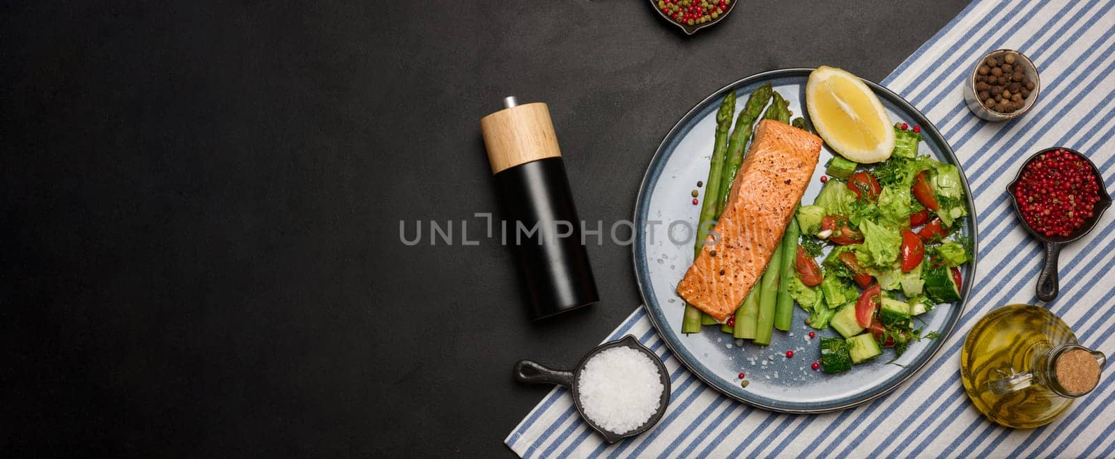 Healthy lunch with grilled trout with asparagus and fresh tomato and cucumber salad, a lemon slice. Food on a blue round plate, top view. Copy space