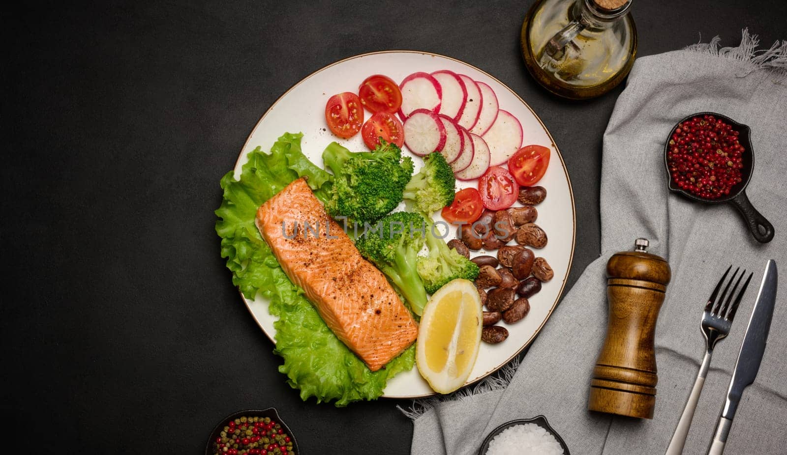 Healthy lunch with grilled salmon on green lettuce, next to vegetables, tomatoes, radishes, broccoli and a portion of beans. Black background