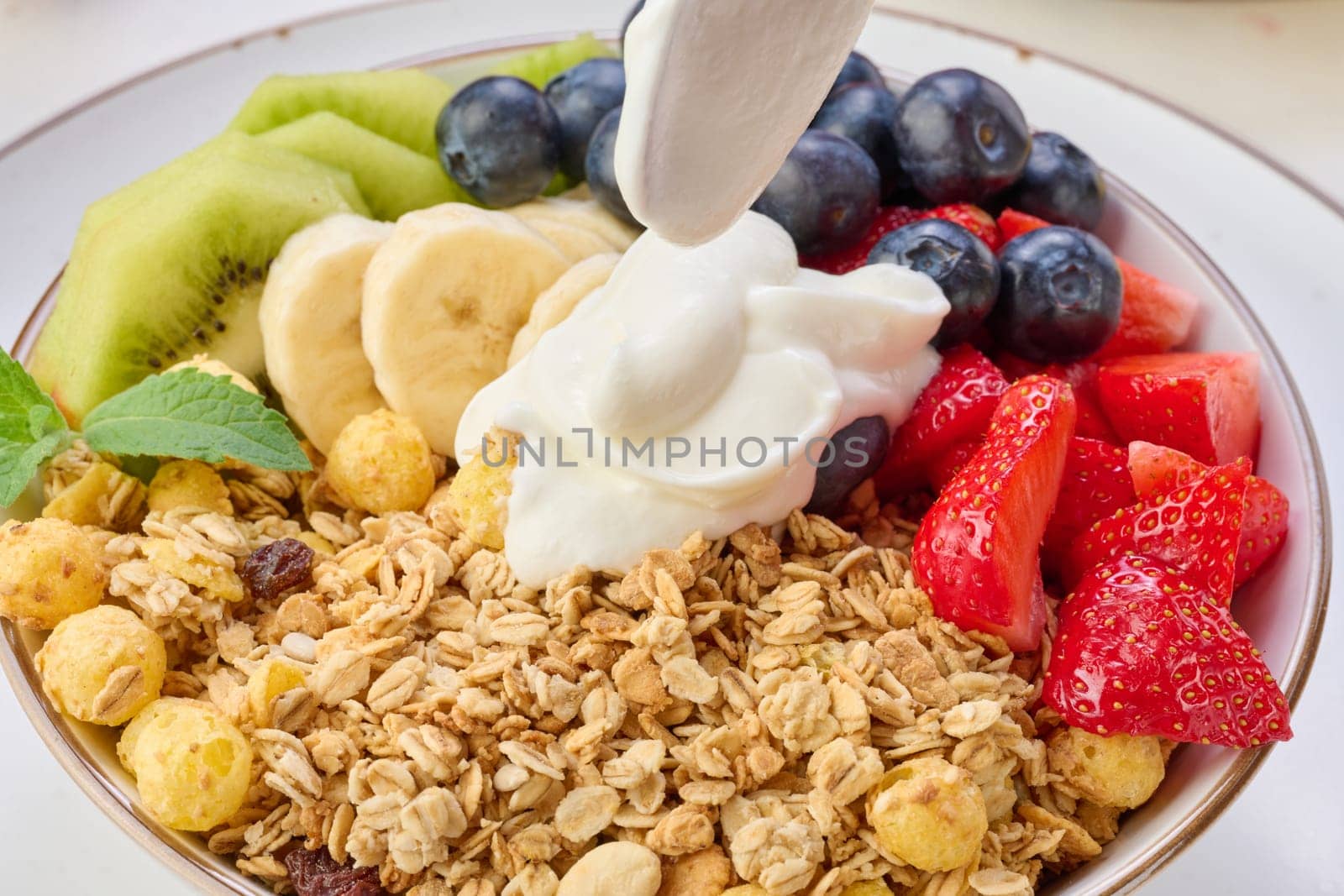 Granola with strawberries, kiwi, banana and blueberries in a round plate topped with yogurt on the table. Healthy and tasty food by ndanko