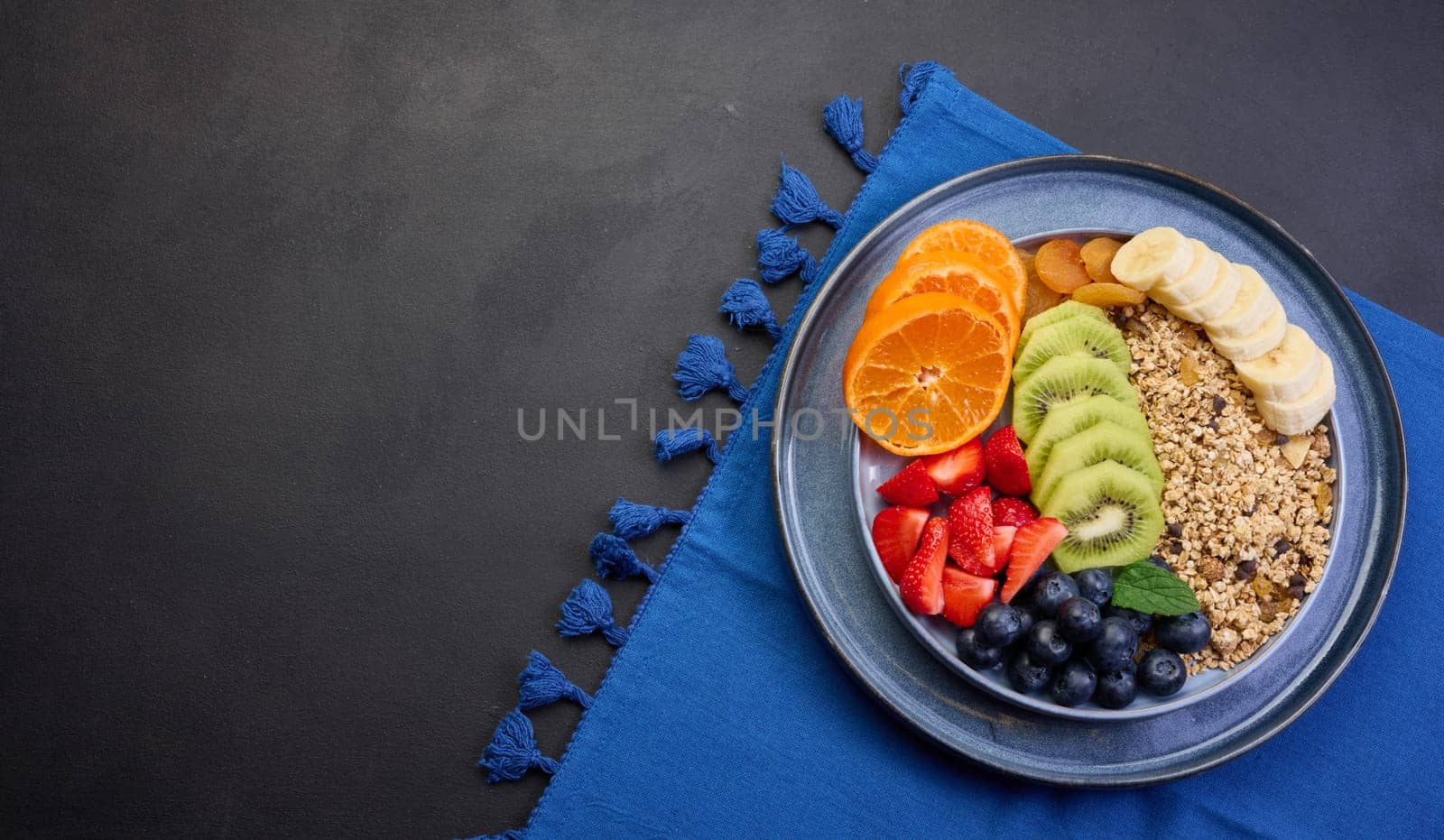 Granola with strawberries, kiwi, banana and blueberries in a round plate on a black table. Healthy and tasty food, top view. Copy space