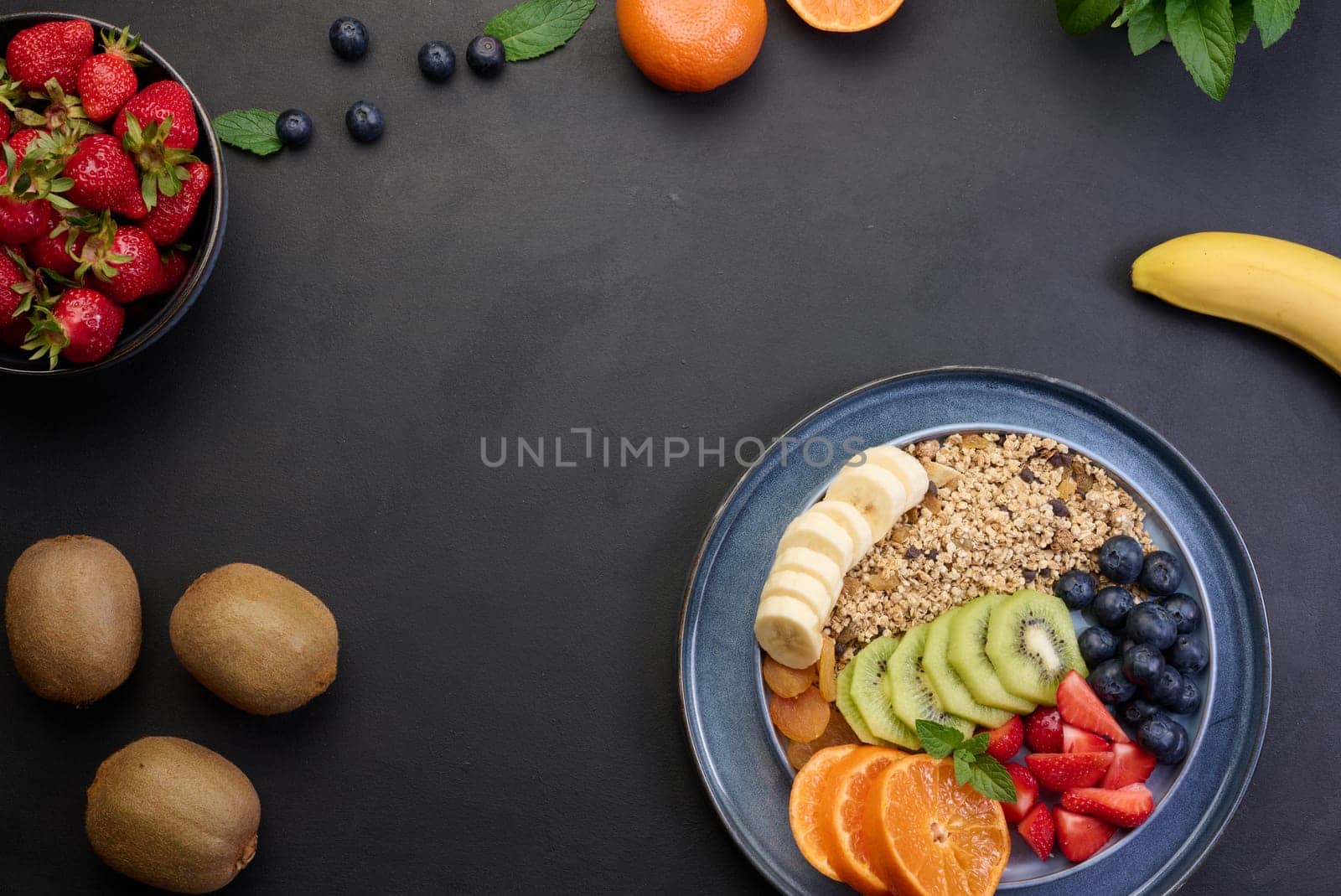 Granola with strawberries, kiwi, banana and blueberries in a round plate on a black table. Healthy and tasty food by ndanko