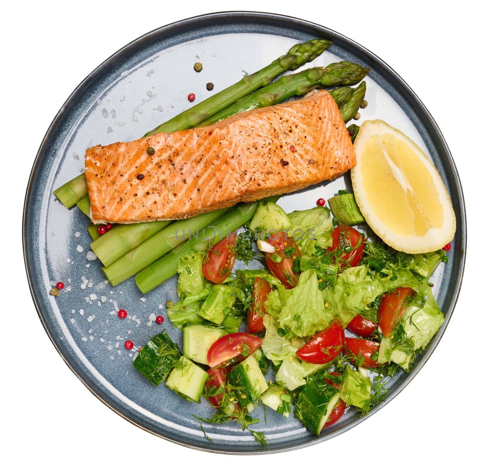 Healthy lunch with grilled trout with asparagus and fresh tomato and cucumber salad, a row of lemon slice. Food on a blue round plate, isolated background