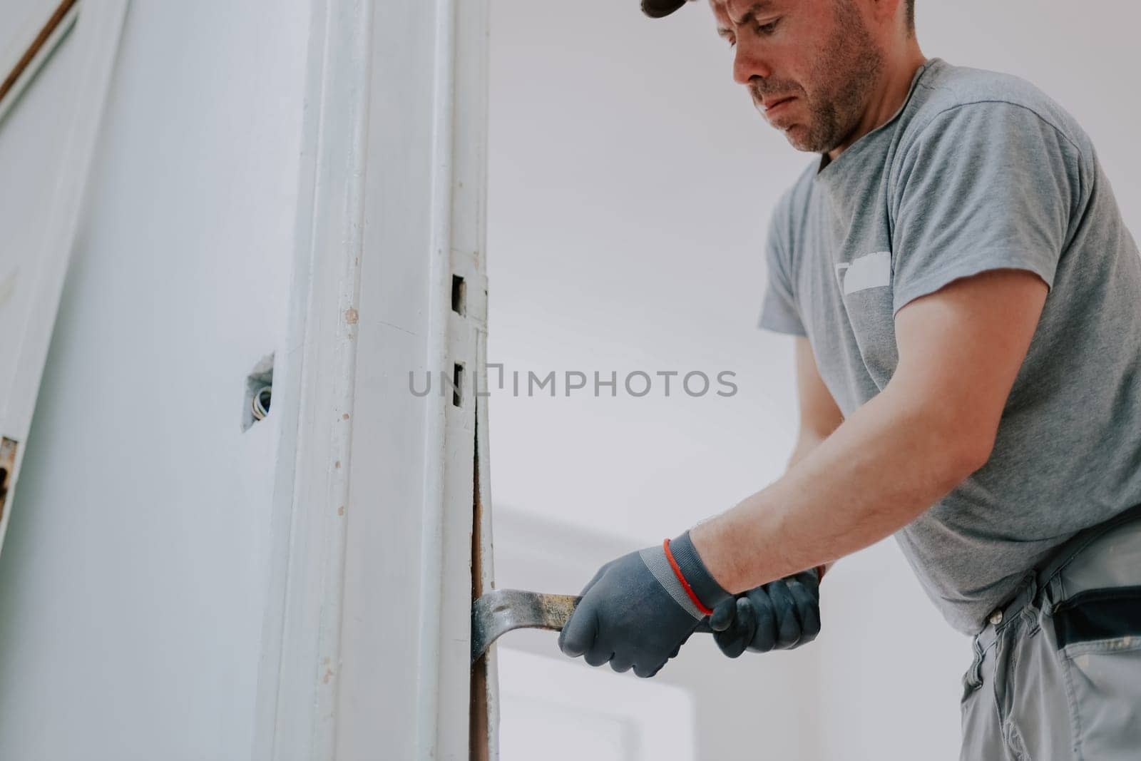 Young caucasian man in uniform and gray textile gloves using a crowbar removes old wooden plinths from a doorway, bottom view close-up with selective focus.Construction work concept.