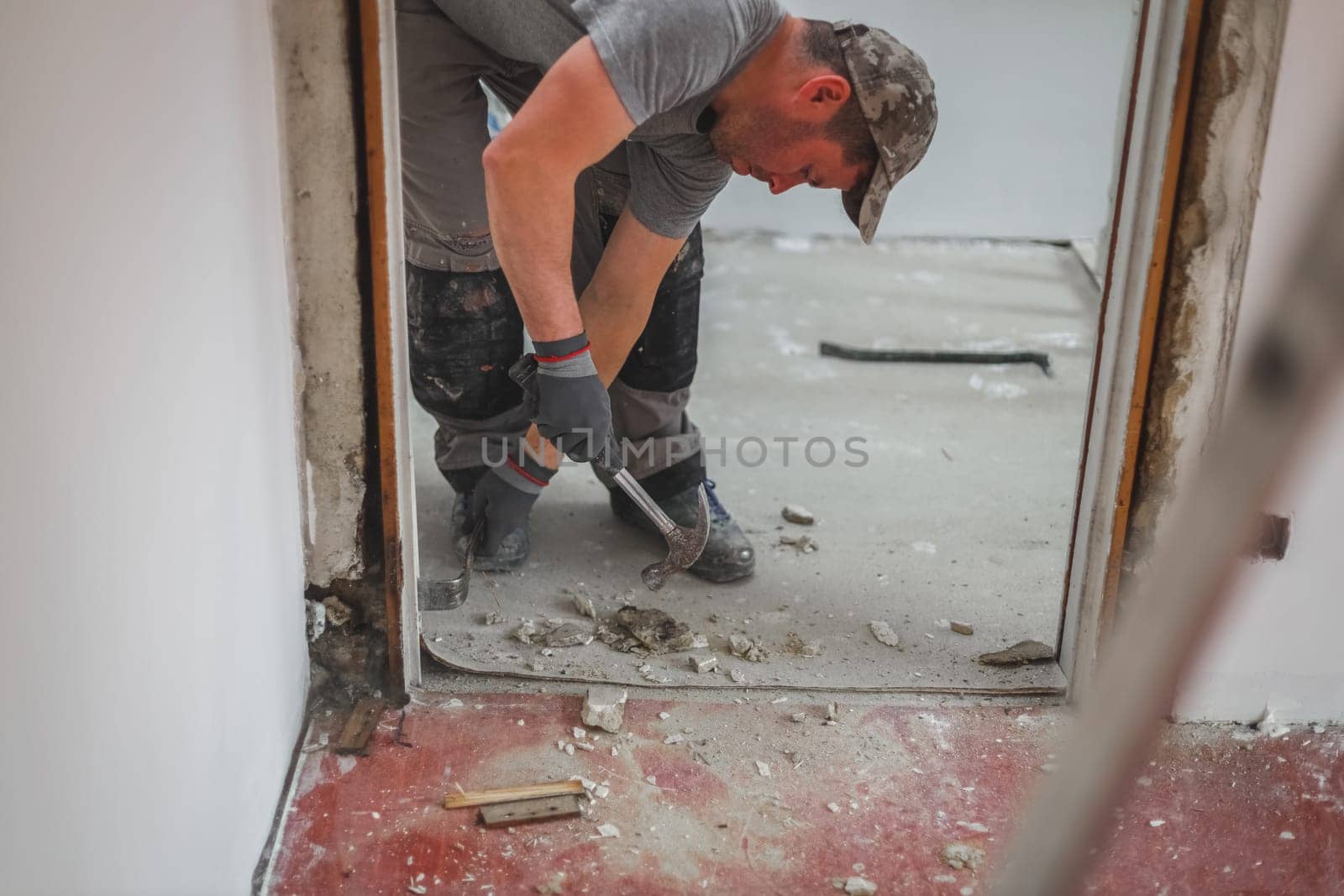 A young caucasian man in a uniform and gray textile gloves with a hammer and crowbar removes old wooden plinths from a doorway while standing in an inclination, close-up view from below with selective focus. Construction work concept.