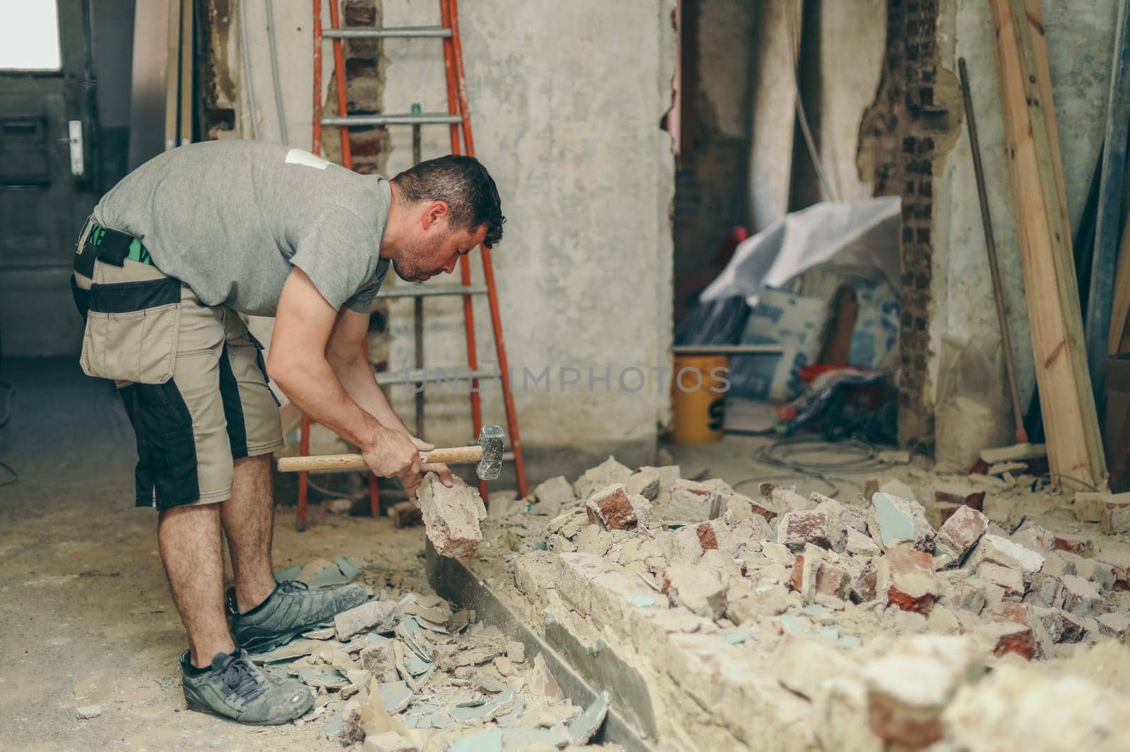 Young caucasian brunette man with a beard in work clothes knocks with a sledgehammer on a dirty brick from a collapsed wall in an old house, close-up side view.Construction work concept.