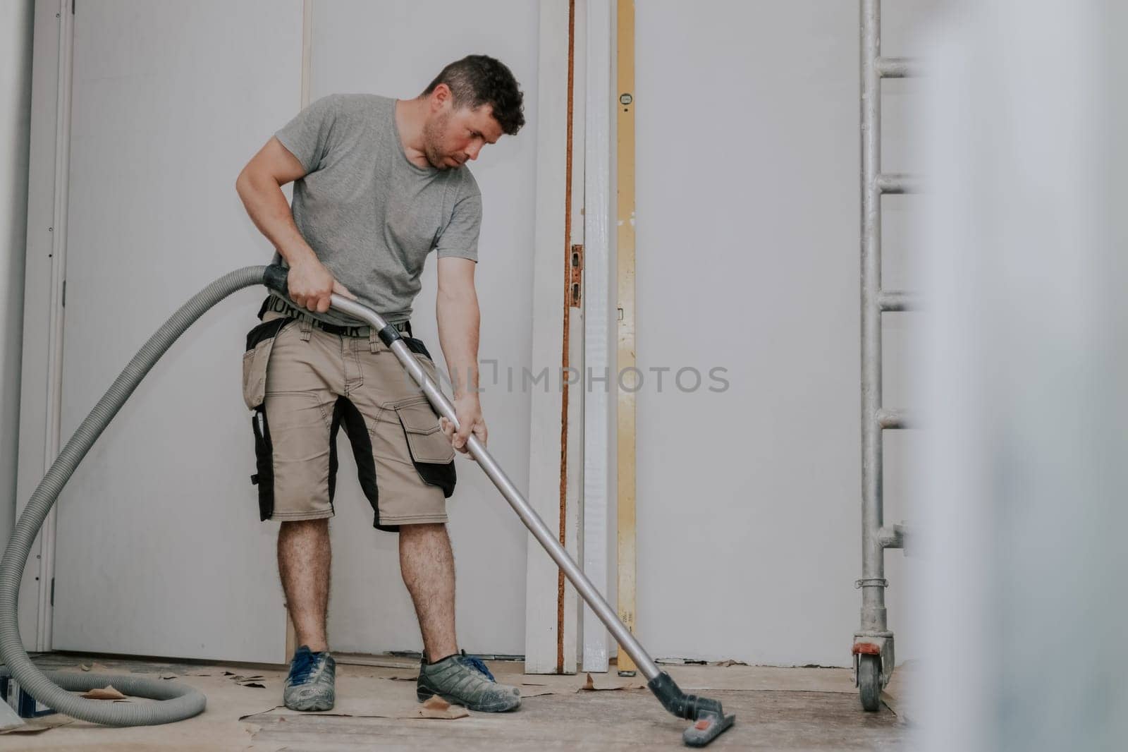 Young caucasian brunette man in work clothes standing sideways vacuums the floor with a construction vacuum cleaner in a room in an old house where renovations are underway, close-up view from below with selective focus.Construction work concept.