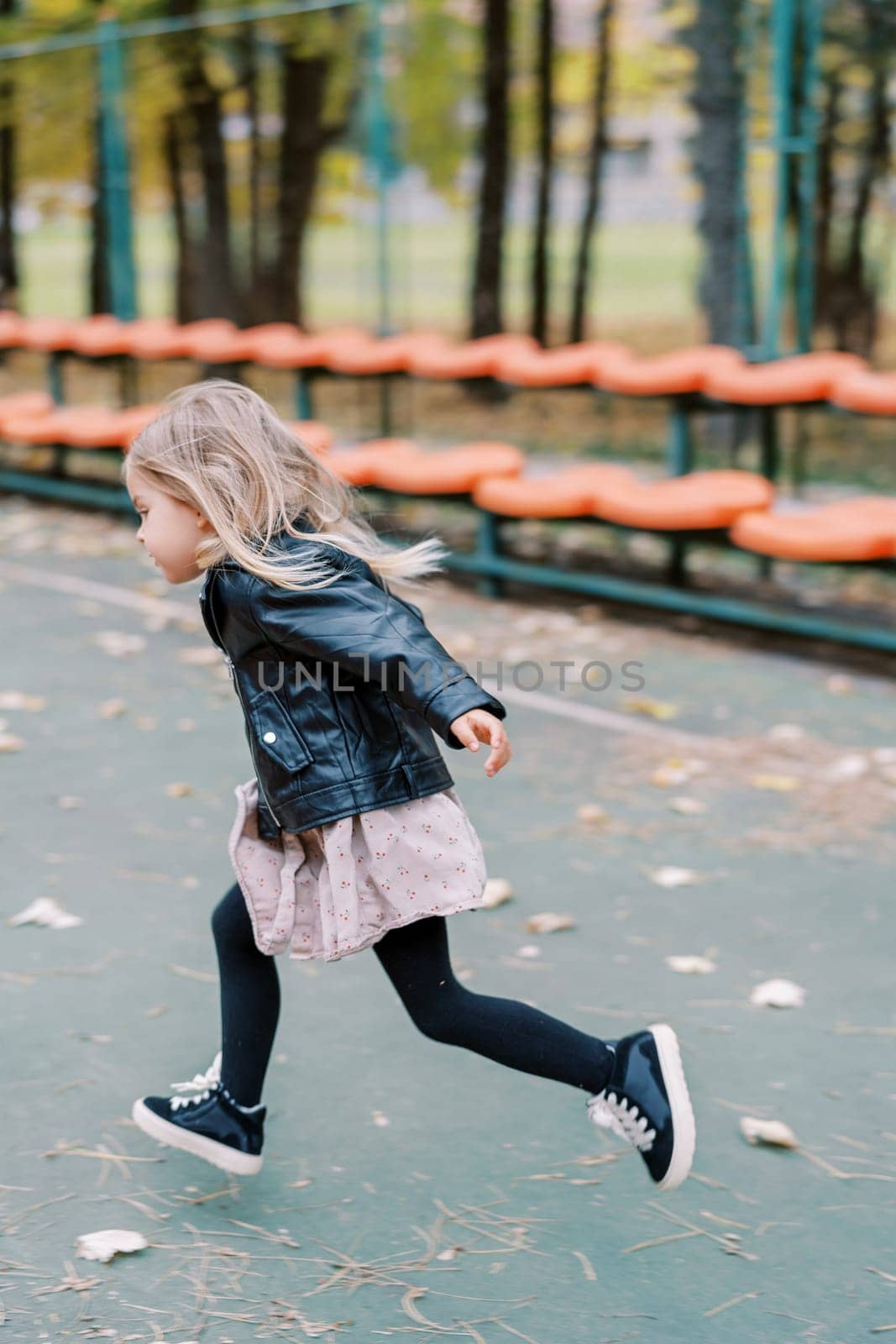 Little girl runs past rows of seats on a sports ground. Side view. High quality photo