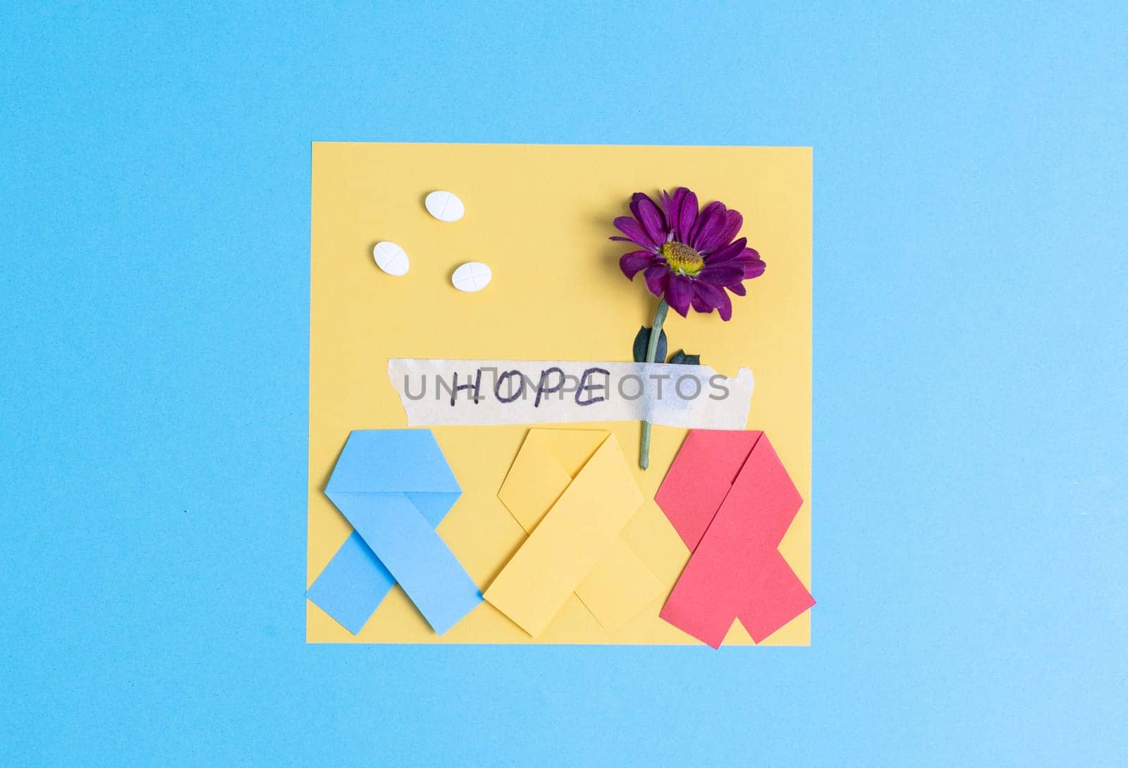 Three multi-colored paper ribbons with a sheet of paper, pills, a gerbera flower and a sticky tape with the inscription hope lie in the center on a blue background with copy space on the sides, flat lay close-up. World Cancer Day concept.