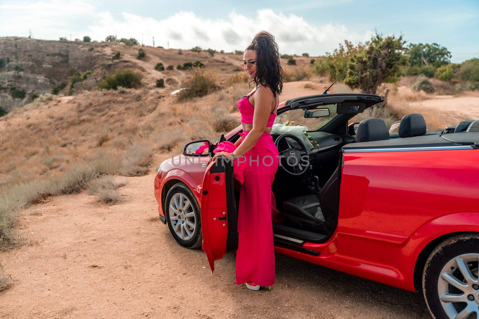 A woman in a red dress stands in front of a red convertible. The scene is set in a desert, with the woman posing for a photo. Scene is playful and fun, as the woman is dressed up. by Matiunina